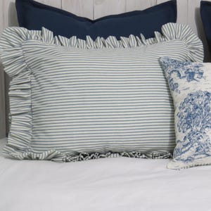 May include: A striped pillow with ruffled edges in blue and white, resting on a white surface. A smaller pillow with a blue and white tree design is visible to the right. A solid navy blue pillow is in the background.