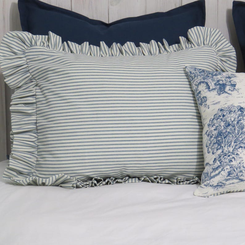 May include: A striped pillow with ruffled edges in blue and white, resting on a white surface. A smaller pillow with a blue and white tree design is visible to the right. A solid navy blue pillow is in the background.