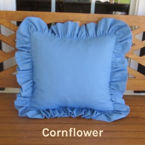 May include: A square, cornflower blue pillow with a ruffled edge. The pillow is centered on a wooden bench. The word "Cornflower" is printed in a cream color at the bottom of the image.