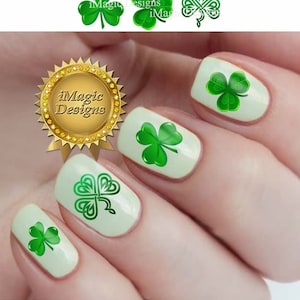 May include: Close-up of a hand with green shamrock nail decals. The nails are painted a light cream color. The shamrocks are in different styles, including a traditional three-leaf clover and a Celtic knot design. The image also includes a gold seal with the text "iMagic Designs".