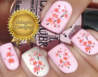 Op de afbeelding: Roze nagelkunst met kersenbloesemdesigns. De nagels zijn geschilderd in lichtroze en wit, met gedetailleerde bloemenstickers. De afbeelding bevat een flesje nagellak en een gouden zegel met de tekst "iMagic Designs".