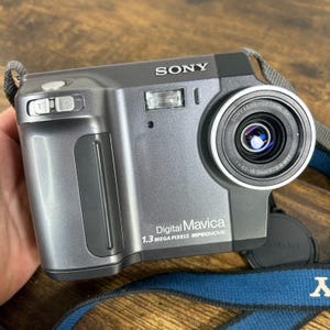 May include: A gray Sony Digital Mavica 1.3 Megapixels MPEGMOVIE camera featuring a Sony Lens Optical 3x with a focal length of 6.1-18.3mm and f/2.8 aperture. The device includes a top-mounted shutter button, a zoom toggle, and a blue neck strap.