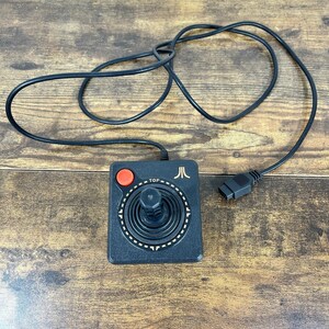 Op de afbeelding: Zwarte Atari-joystick met een rode knop en een zwarte kabel. De joystick heeft een cirkelvormig ontwerp rond de stick en het Atari-logo. Het woord "TOP" staat op de joystick.
