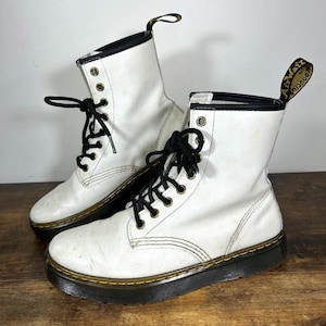 May include: A pair of white leather boots with black laces and a black sole. The boots have a mid-calf height and feature a yellow stitch detail around the sole. The boots are on a wooden surface.