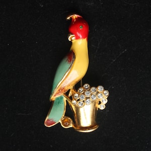 May include: A gold-tone brooch featuring a yellow, green, and red bird perched on a gold bucket filled with clear rhinestones.