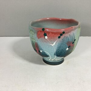 May include: A hand-painted ceramic bowl with a unique floral design. The bowl features a red rim, with blue, green, and white accents. The exterior is decorated with abstract floral patterns. The bowl has a dark green base.