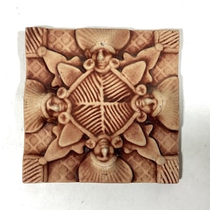 May include: Square, tan-colored tile with a raised, symmetrical design. The central motif is a diamond shape with a leaf-like pattern, surrounded by stylized faces and floral elements. The tile has a textured surface.