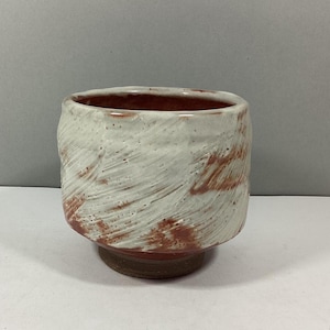 May include: A white ceramic cup with a red glaze and a textured surface. The cup is round and has a wide mouth.