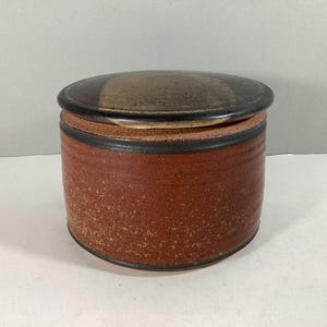 May include: A round, ceramic jar with a lid. The jar is a reddish-brown color with a textured surface. The lid is a darker brown with a black rim. The jar is likely used for storage.