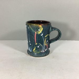May include: A handmade ceramic mug with a dark teal base and handle. The mug features a dark brown interior and is decorated with yellow, red, and white floral designs. The mug is approximately 4 inches tall.