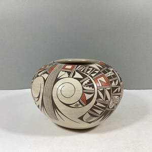 May include: A round, off-white ceramic pot with a wide opening. The pot is decorated with a repeating geometric pattern in brown and red. A large spiral design is on the front of the pot.