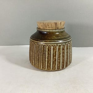 May include: A ceramic jar with a cork lid. The jar has a brown and tan color scheme with vertical ribbed detailing on the body. The jar is a small, decorative storage container.