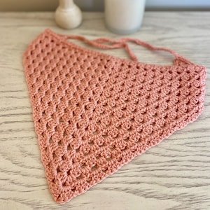 A peach crocheted bandana lays on a dresser. This boho style hair scarf has a delicate open stitch pattern.