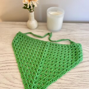 A green crocheted bandana lays on a dresser. This boho style hair scarf has a delicate open stitch pattern. There are ties on the bandana to secure to the hair and head.
