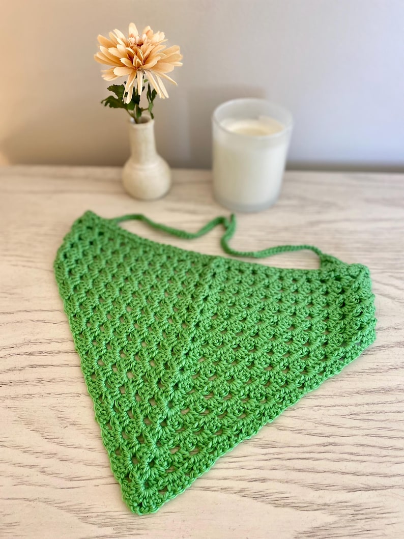 A green crocheted bandana lays on a dresser. This boho style hair scarf has a delicate open stitch pattern. There are ties on the bandana to secure to the hair and head.
