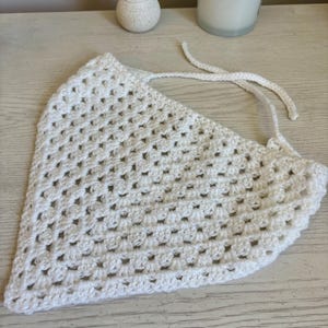May include: A white triangular bandana crafted with a crochet granny square stitch pattern. The accessory features a repeating openwork design and includes two thin crocheted ties extending from the top corners for securing the garment in place.