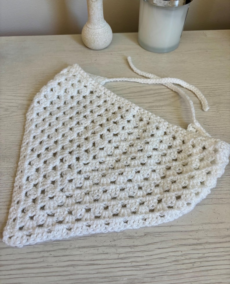 May include: A white triangular bandana crafted with a crochet granny square stitch pattern. The accessory features a repeating openwork design and includes two thin crocheted ties extending from the top corners for securing the garment in place.