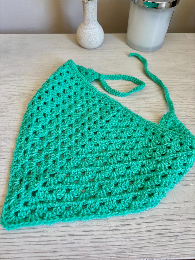 May include: A bright green triangular crochet bandana featuring a classic granny square stitch pattern. The accessory is crafted from yarn and includes two long, thin crocheted ties at the top corners for securing the piece around the head or neck.