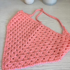 May include: A triangular crochet bandana in a solid peach color, featuring a classic granny square stitch pattern with openwork clusters and two long, thin crocheted ties extending from the top corners for securing the garment.