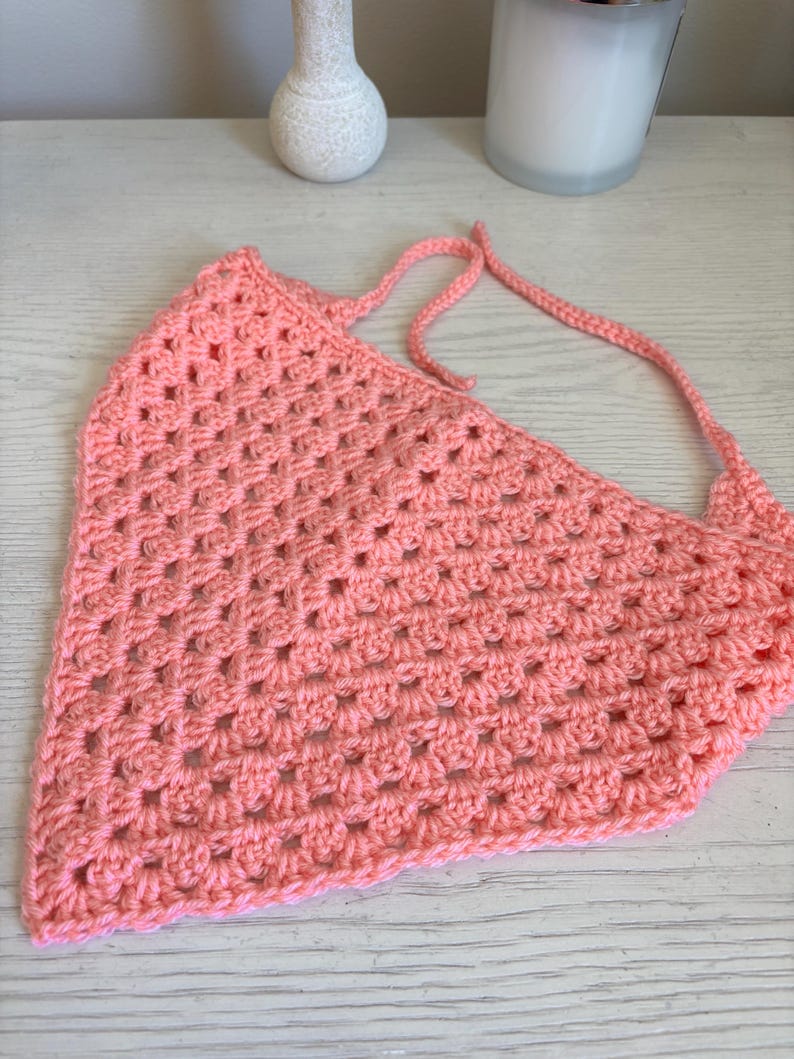 May include: A triangular crochet bandana in a solid peach color, featuring a classic granny square stitch pattern with openwork clusters and two long, thin crocheted ties extending from the top corners for securing the garment.
