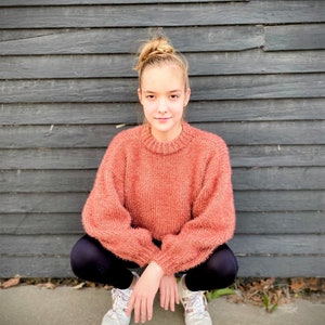 May include: A young person wearing a peach-colored fuzzy sweater, black leggings, and white sneakers. They are sitting with their legs crossed in front of a gray wooden wall.
