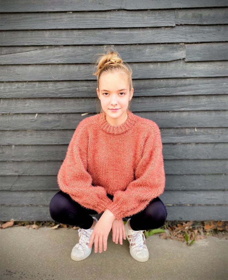 May include: A young person wearing a peach-colored fuzzy sweater, black leggings, and white sneakers. They are sitting with their legs crossed in front of a gray wooden wall.