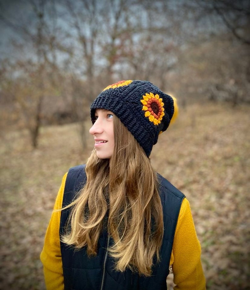 May include: A black crocheted beanie with a sunflower design. The beanie has a pompom on top and is worn by a person with long blonde hair.
