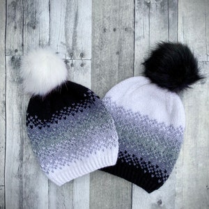 May include: Two knitted winter hats with pom poms. One hat is black with white and grey stripes and a white pom pom. The other hat is white with black and grey stripes and a black pom pom.