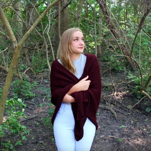 Puede incluir: Una joven con el pelo largo y rubio lleva una chalina de punto marr&oacute;n y pantalones blancos. Est&aacute; de pie en una zona arbolada con vegetaci&oacute;n verde y ramas marrones.