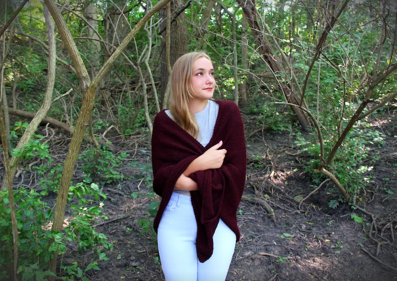 Puede incluir: Una joven con el pelo largo y rubio lleva una chalina de punto marr&oacute;n y pantalones blancos. Est&aacute; de pie en una zona arbolada con vegetaci&oacute;n verde y ramas marrones.
