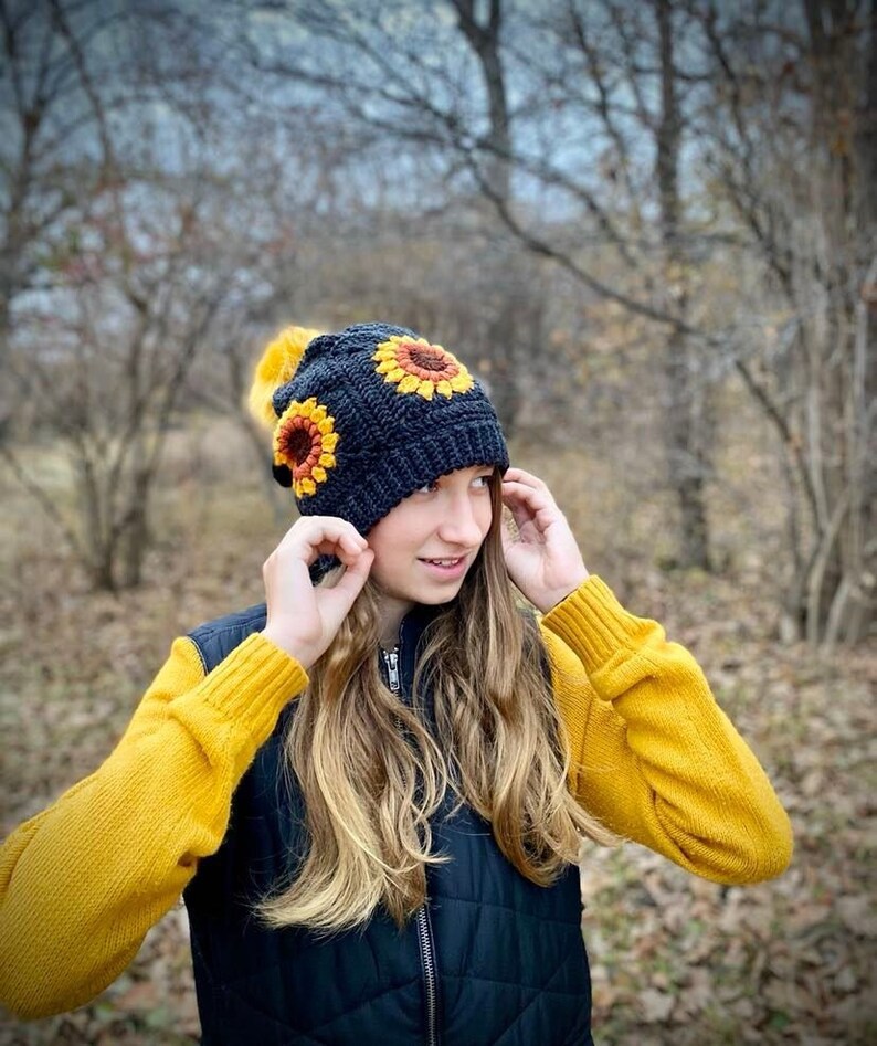 May include: A black crocheted beanie with a pom pom on top and three crocheted sunflower designs. The beanie is being worn by a person with long blonde hair.