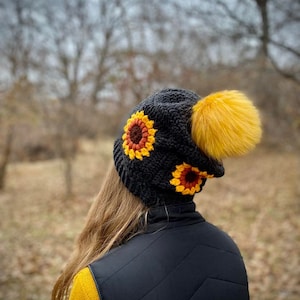 May include: A black crochet hat with a yellow pom pom and two sunflower appliques. The hat is worn by a person with long brown hair and a yellow sweater.