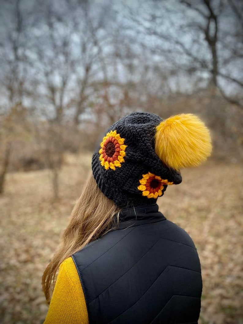 May include: A black crochet hat with a yellow pom pom and two sunflower appliques. The hat is worn by a person with long brown hair and a yellow sweater.