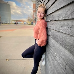 May include: A young person wearing a pink fuzzy sweater and black leggings stands against a gray wooden wall. They are wearing white sneakers with black stripes.