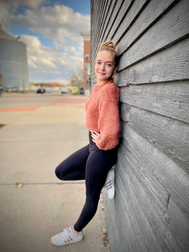 May include: A young person wearing a pink fuzzy sweater and black leggings stands against a gray wooden wall. They are wearing white sneakers with black stripes.