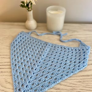A light blue crocheted bandana lays on a dresser. This boho style hair scarf has a delicate open stitch pattern. There are ties on the bandana to secure to the hair and head.