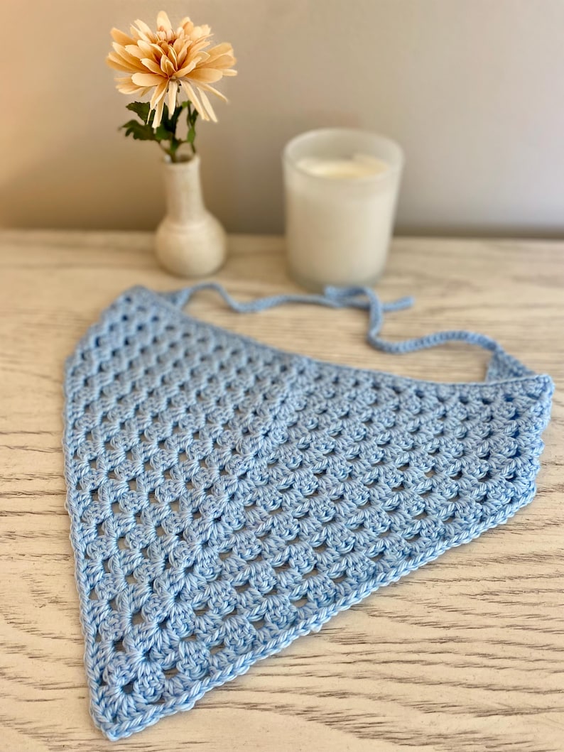 A light blue crocheted bandana lays on a dresser. This boho style hair scarf has a delicate open stitch pattern. There are ties on the bandana to secure to the hair and head.