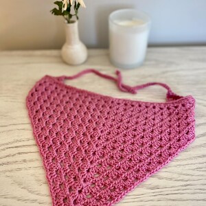 A pink crocheted bandana lays on a dresser. This boho style hair scarf has a delicate open stitch pattern. There are ties on the bandana to secure to the hair and head.