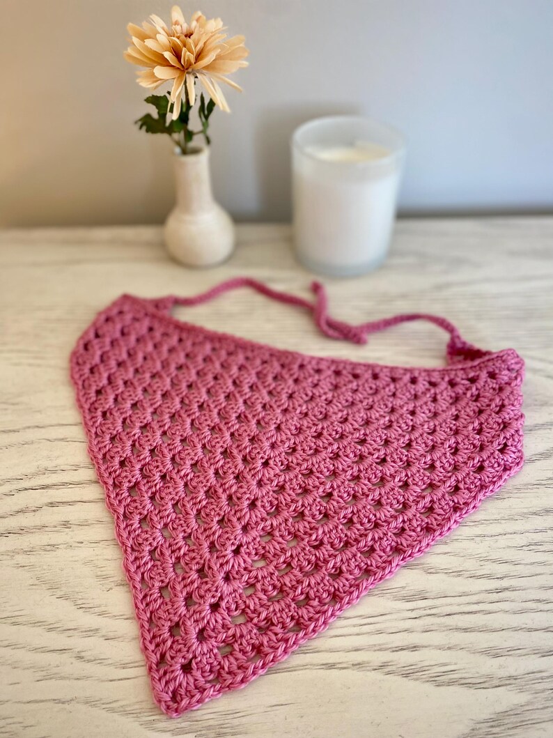 A pink crocheted bandana lays on a dresser. This boho style hair scarf has a delicate open stitch pattern. There are ties on the bandana to secure to the hair and head.