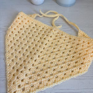 May include: A pale yellow triangular bandana crafted with a crochet granny square stitch pattern. The accessory features a textured, open-work design and includes two long, thin crocheted ties at the top corners for securing the item in place.