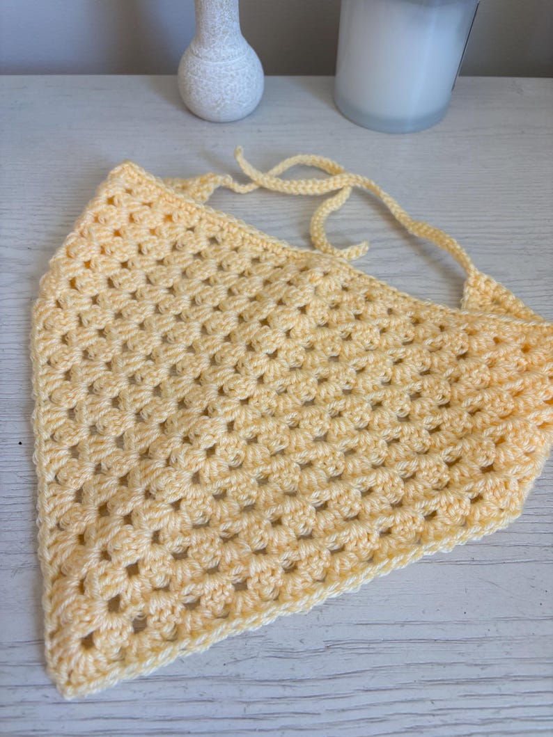 May include: A pale yellow triangular bandana crafted with a crochet granny square stitch pattern. The accessory features a textured, open-work design and includes two long, thin crocheted ties at the top corners for securing the item in place.