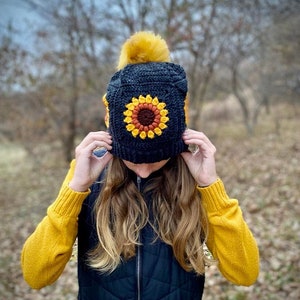 May include: A black crochet beanie with a yellow pom pom and a crocheted sunflower design. The beanie is being worn by a person with long blonde hair.