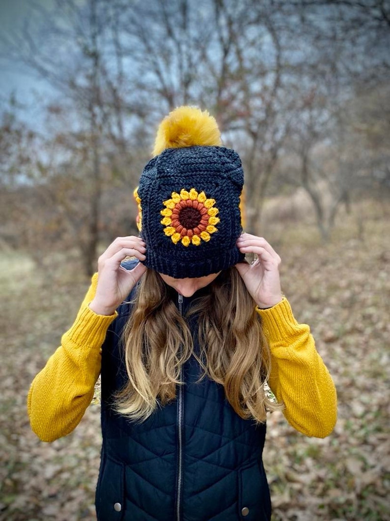 May include: A black crochet beanie with a yellow pom pom and a crocheted sunflower design. The beanie is being worn by a person with long blonde hair.