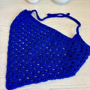 May include: A vibrant royal blue triangular bandana crafted with a crochet granny square stitch pattern. The accessory features an open, airy texture throughout the fabric and includes two long, thin crocheted ties at the top corners for securing.