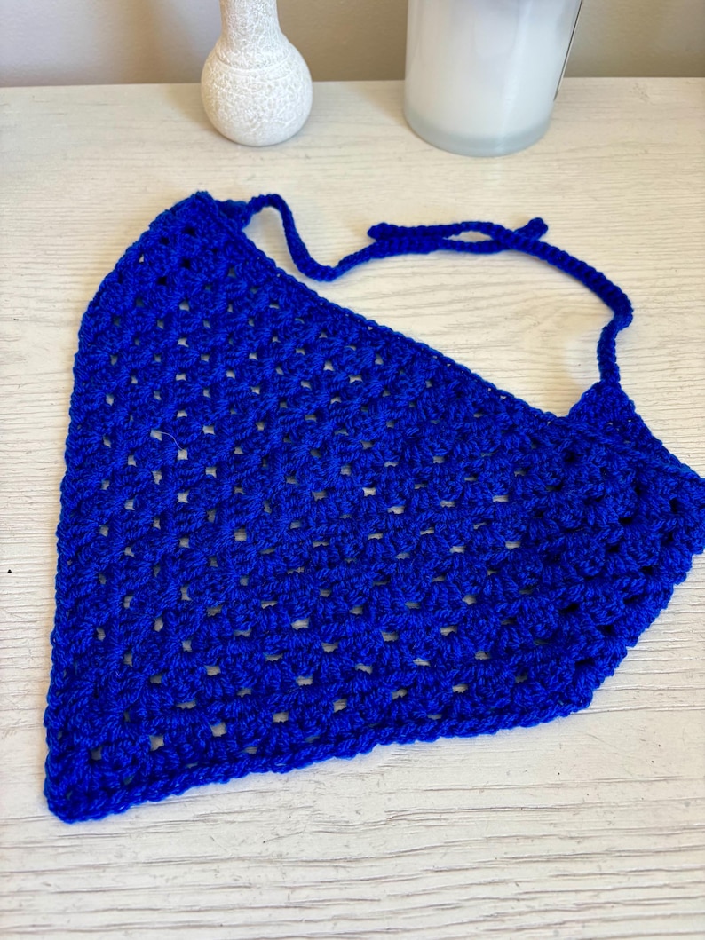 May include: A vibrant royal blue triangular bandana crafted with a crochet granny square stitch pattern. The accessory features an open, airy texture throughout the fabric and includes two long, thin crocheted ties at the top corners for securing.