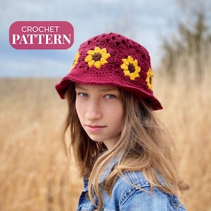 以下が含まれることがあります： 赤ワイン色のベースに黄色のひまわりがあしらわれた、かぎ針編みのバケットハット。長い茶色の髪の女性が被っています。ピンク色の長方形に白い文字で「CROCHET PATTERN」と書かれています。