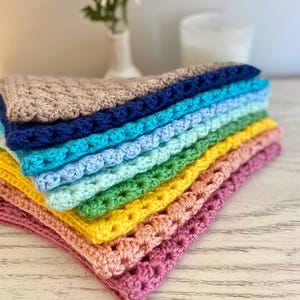 A stack of crocheted bandanas in various colors, including pink, yellow, green, blue, and brown sit on a dresser. The crocheted bandanas are folded in half.