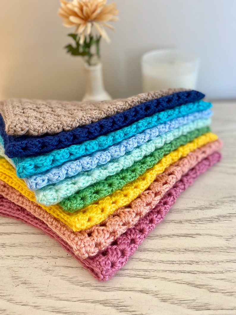 A stack of crocheted bandanas in various colors, including pink, yellow, green, blue, and brown sit on a dresser. The crocheted bandanas are folded in half.