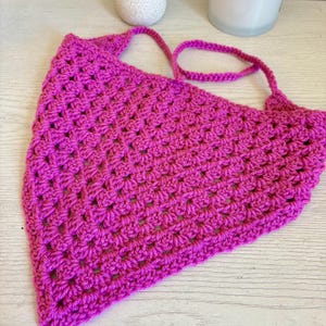 May include: A bright magenta triangular bandana crafted with a textured crochet stitch pattern. The accessory features a pointed bottom edge and two thin crocheted ties extending from the top corners for securing the garment in place.