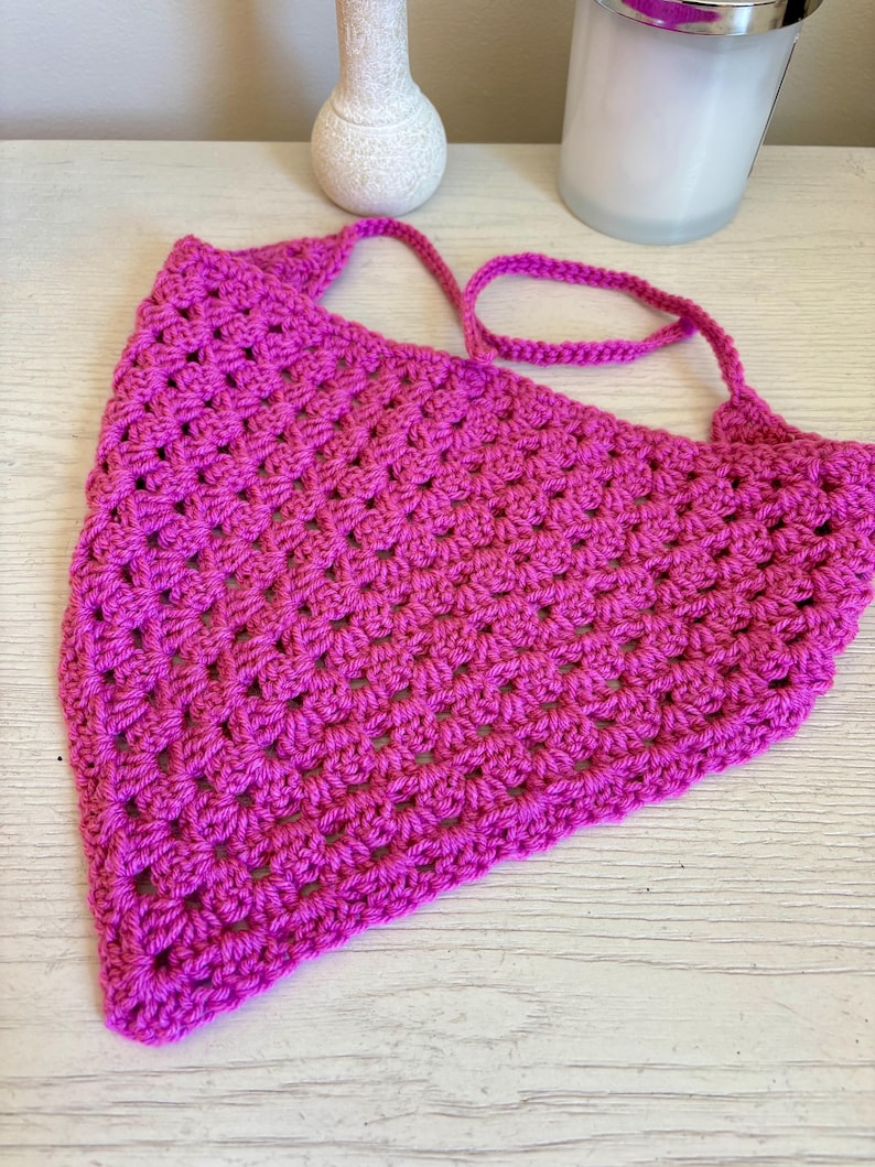 May include: A bright magenta triangular bandana crafted with a textured crochet stitch pattern. The accessory features a pointed bottom edge and two thin crocheted ties extending from the top corners for securing the garment in place.